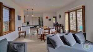 A modern living room with a grey sofa, a dining area with a table and chairs, a kitchen with a fridge and stove, and large windows with a stone wall view.