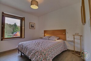 Un dormitorio con cabecera de madera, cama con ropa de cama floral, mesita de noche, lámpara colgante y ventana con vistas a un coche aparcado y vegetación exterior.