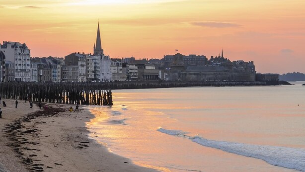 A coastal cityscape at sunset with a prominent church spire, a promenade, and a row of buildings along the waterfront.