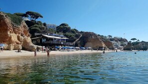 A coastal beach with a building that has a terrace and umbrellas. - Charming Sea View Flat w/Pool, 20 meters from the Beach (Albufeira)