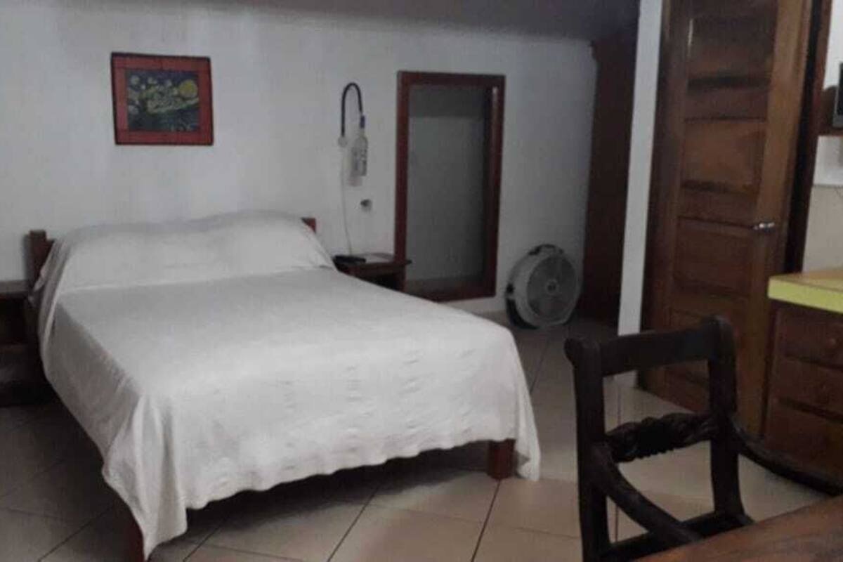 Quarto com cama, mesa de cabeceira, cadeira, ventilador e porta.
