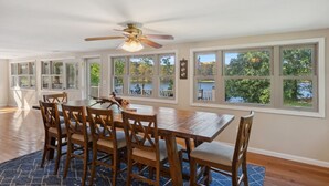 A dining area with a wooden table and chairs, large windows with a view of trees and a body of water, and a ceiling fan.