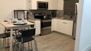 Una cocina compacta con microondas, estufa, horno y lavadero. Hay una mesa de comedor con sillas y algunos objetos encima.