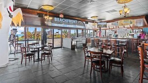 En restaurantindretning med strandtema, inklusive et skilt der står 'BEACHFRONT Kitchen & Bar', og udsigt til palmer udenfor.