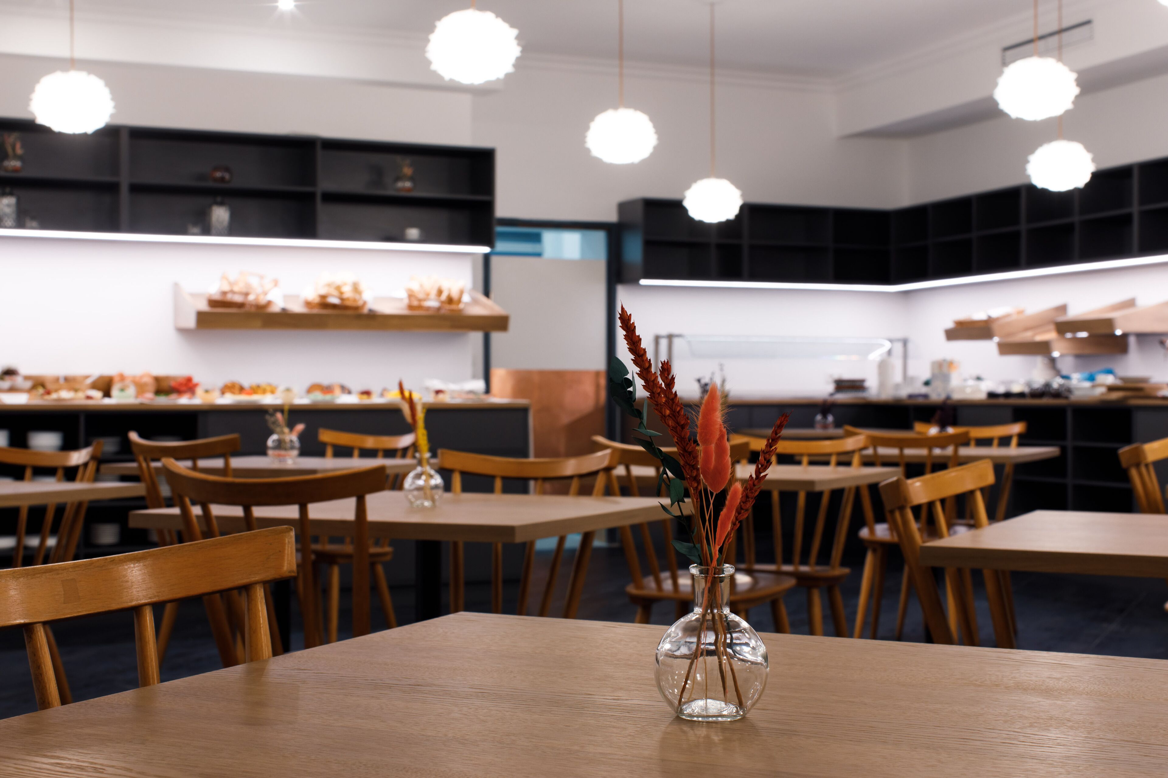 Un espace de restauration moderne avec des tables et des chaises en bois, un vase central agrémenté d’éléments décoratifs et un éclairage suspendu.