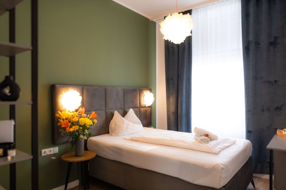 A neatly made bed with white linens, a bedside table with a vase of flowers, and a modern chandelier.