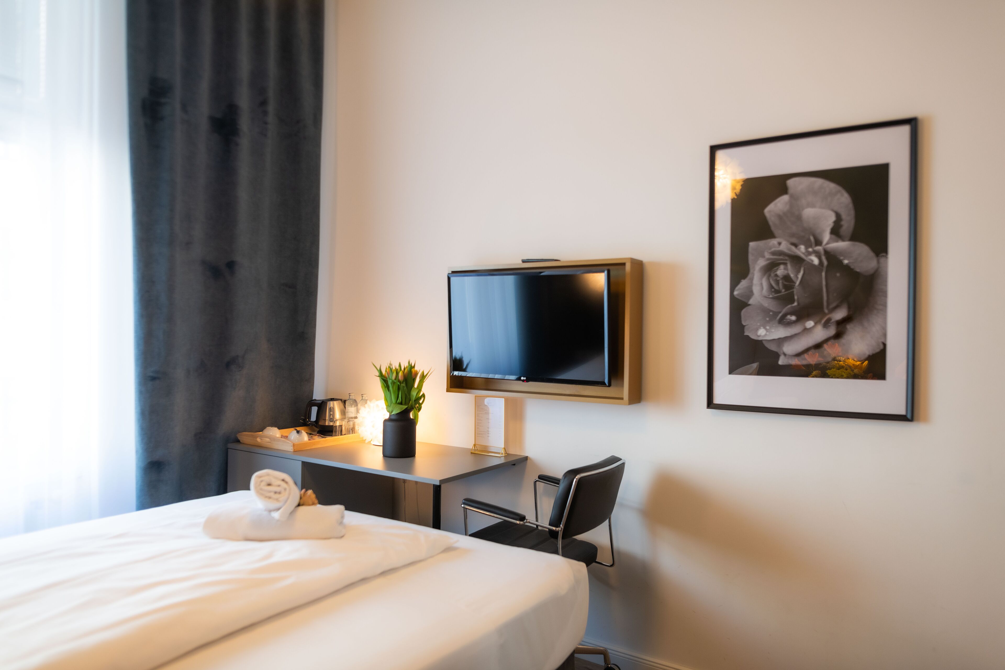 Ein Hotelzimmer mit einem Bett, einem Schreibtisch mit Fernseher, einem Sessel, einer Zimmerpflanze und einem gerahmten Bild an der Wand.
