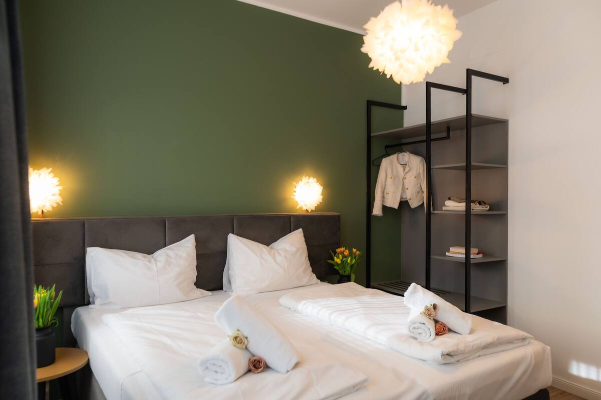 A hotel room with a bed, two pillows, a bedside table with a plant, a nightstand, a wall-mounted shelf with towels and a robe, and a pendant light.