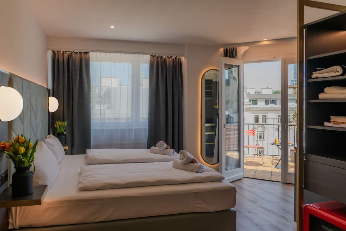 A hotel room with two beds, a large window with curtains, a view of the city, and a balcony with a table and chairs.