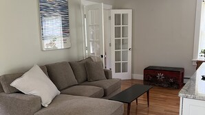 Un salon comprenant un canapé d’angle, une table basse, un plancher en bois, un meuble blanc et une porte à panneaux de verre.