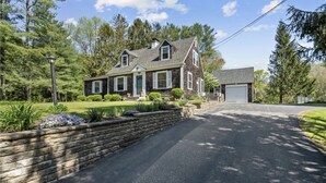 Une maison à deux étages avec un garage, entourée d’un mur de soutènement en pierre et d’une végétation luxuriante.