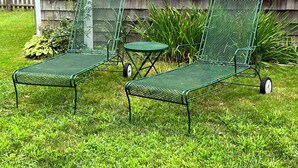 Deux chaises de jardin en métal vert sont placées devant une maison en bois dotée d’une fenêtre.