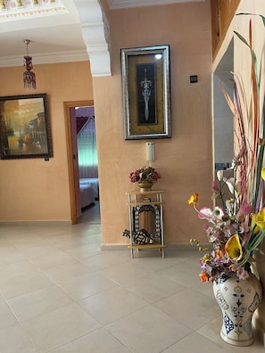 Un couloir avec un sol carrelé, un tableau encadré, un vase rempli de fleurs et un vase décoratif contenant des arums.