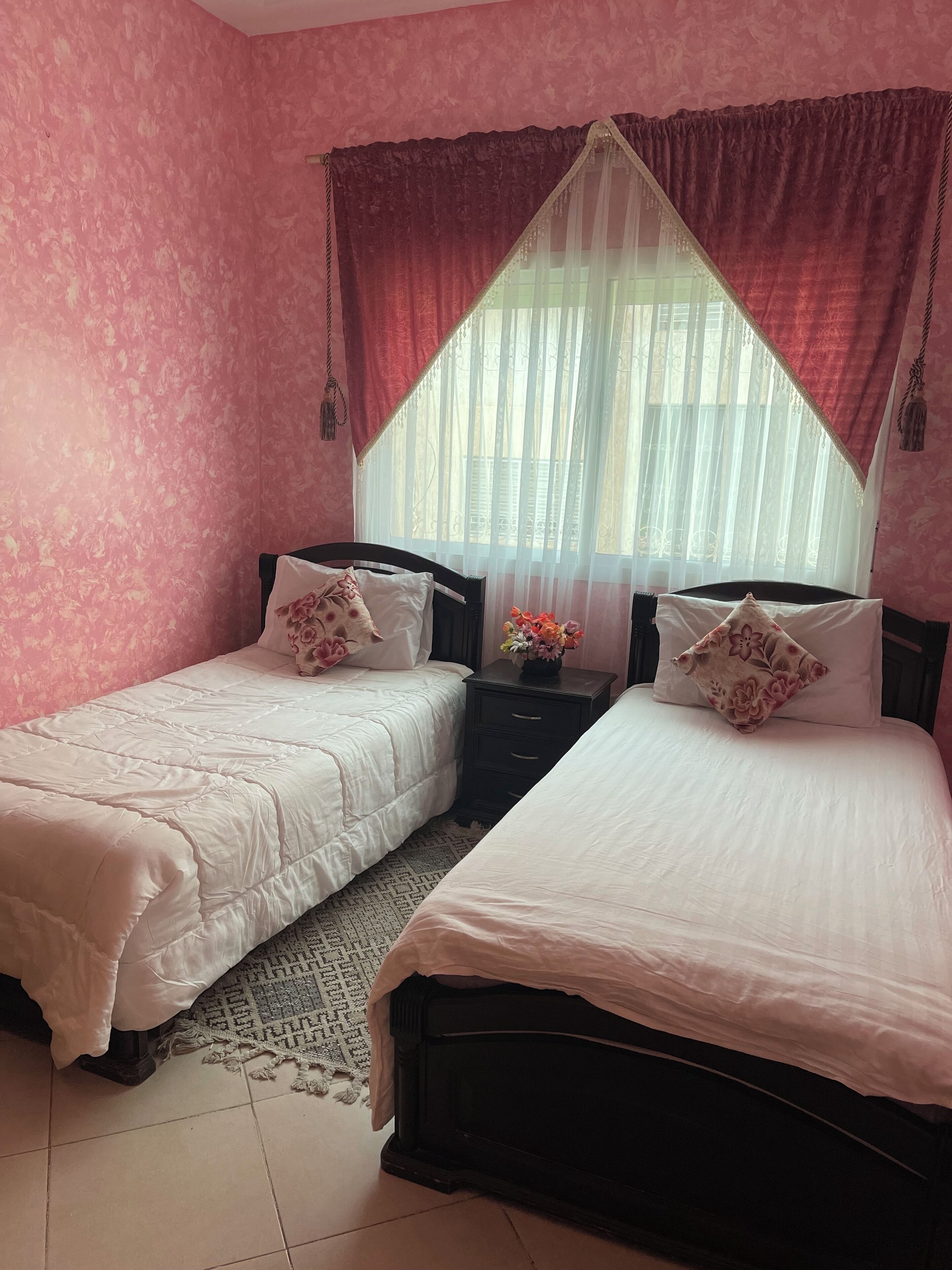 Un dormitorio con dos camas, una mesita de noche y una ventana con cortinas traslúcidas.