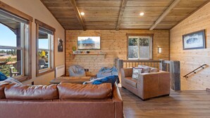 A cozy cabin interior with a wooden ceiling, a brown leather sofa, a cushioned armchair, and a large window offering a view of the outdoors.