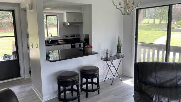 A modern kitchen with a central island, two stools, a small table with a plant, and a view of a garden through large windows.