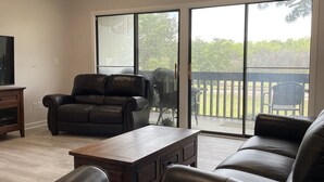 A spacious living room with a large sliding glass door leading to a deck, a ceiling fan, and a flat-screen TV mounted on the wall.