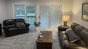 A living room with a black leather sofa, a wooden coffee table, and a lamp. There is a sliding glass door leading to a balcony.