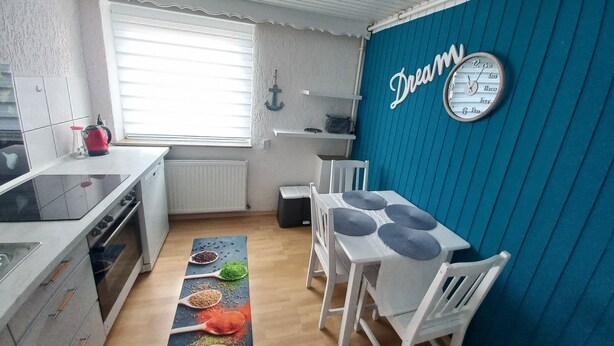 Een kleine keuken met een raam, witte kastjes, een spoelbak en een eethoek met een tafel en stoelen.