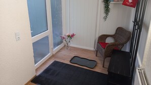Een kamer met een schuifdeur, een rieten stoel met een rode kussen, een zwarte mat, een EHBO-kit, en een plankje met een cactus en een 'hello' bordje.