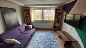 Een woonkamer met een paarse bank, een vloerkleed, een houten bankje en een boekenkast.