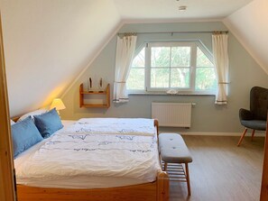 Ein Schlafzimmer mit einem Bett, einer Bank, einem Sessel, einem Fenster mit Vorhängen, einem Regal mit Dekoration und einem Heizkörper.