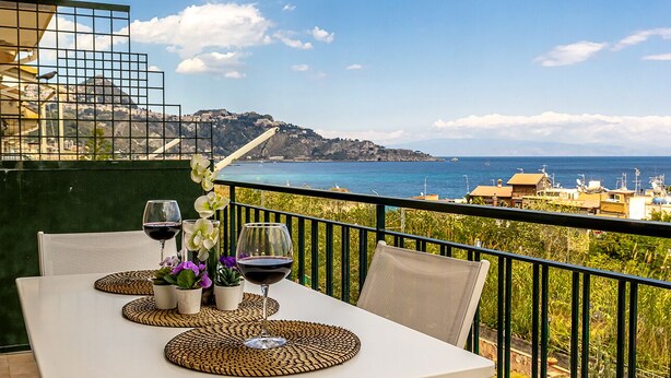 Un balcón con una mesa dispuesta para dos personas, que incluye una copa de vino y flores, con vistas a un paisaje costero con edificios y una colina.