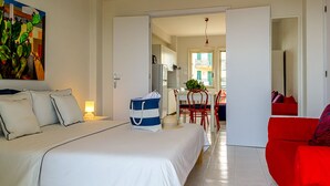 Habitación de hotel con cama, sofá rojo, zona de comedor y cocina visible a través de una puerta abierta.