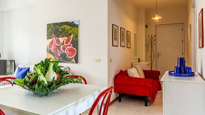 Un comedor con una mesa, sillas y un frutero. Hay un sofá rojo y un armario blanco con un objeto azul encima.