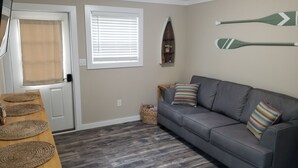 Een woonkamer met een grijze bank, twee geweven onderzetters op een houten tafel, een mand en een surfboarddecoratie aan de muur.