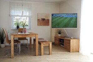 Una sala de estar con una mesa de comedor de madera, sillas, un banco, un televisor montado en la pared, una ventana con vista a follaje, y una planta en maceta.