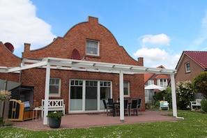 A brick house with a white pergola patio, a covered outdoor seating area, and a garden with flowers.