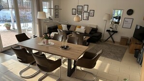 A modern living room with a wooden dining table, brown chairs, a black sofa, and a large window providing a view of the outdoors.