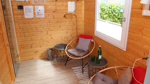A wooden cabin interior with a sauna poster, a chair, a small table with a bottle, and a window with a view of greenery.