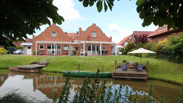 A riverside property with a brick house, a dock with boats, and a deck with outdoor seating.