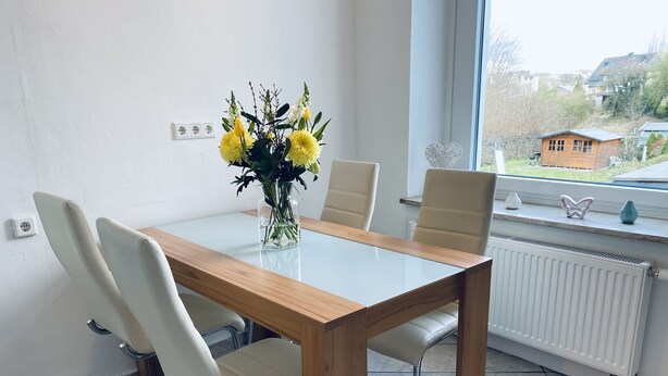 Un espace repas moderne avec une table en verre, quatre chaises blanches et un vase rempli de fleurs jaunes.