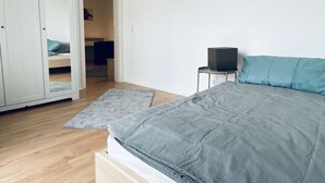 Une chambre à coucher comprenant un lit, une armoire, une petite table avec un haut-parleur, et une porte donnant sur une autre pièce.
