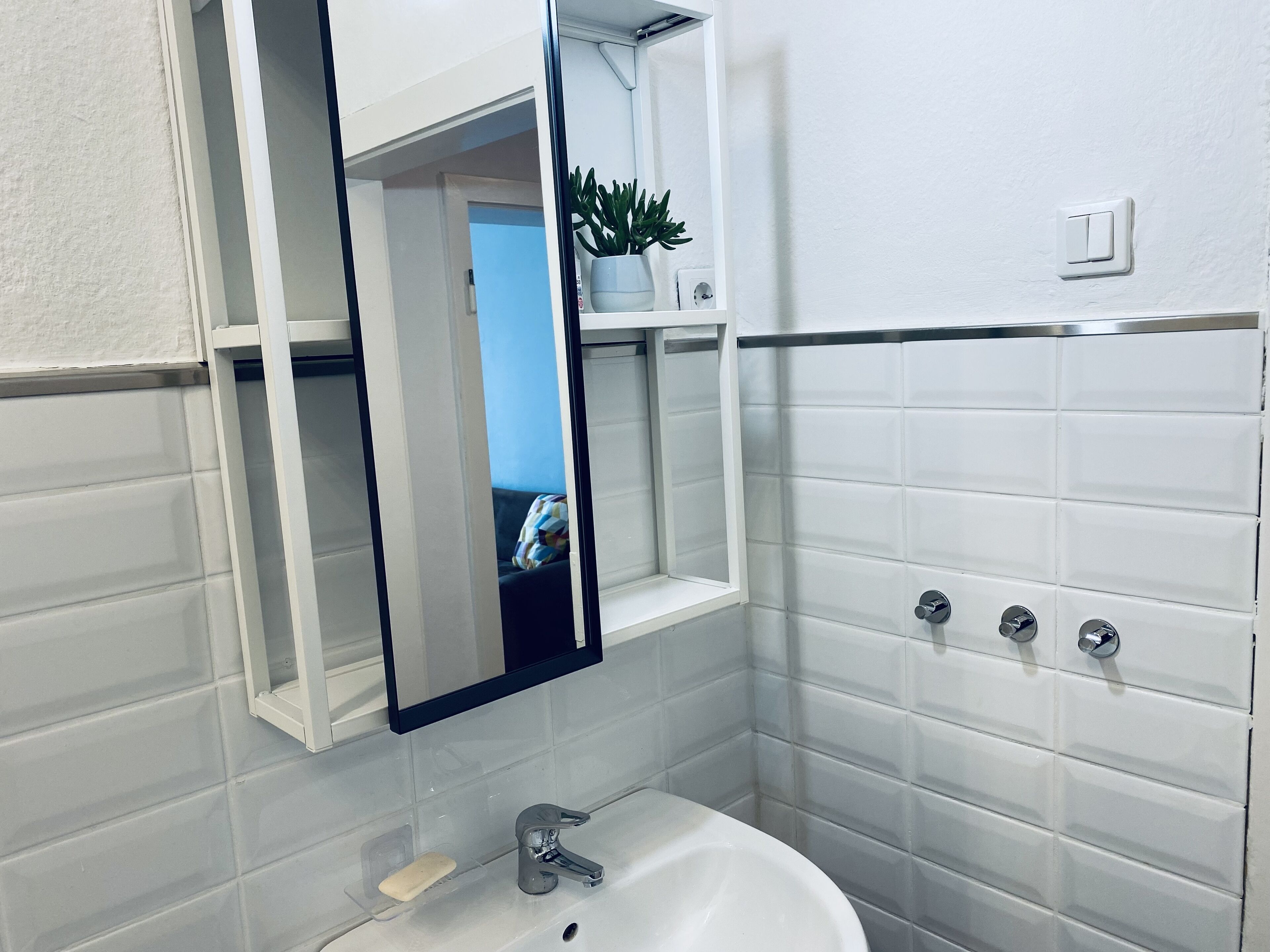 Un baño con paredes de azulejos blancos, un lavamanos blanco, un espejo y una planta en maceta sobre una estantería.