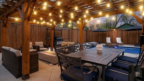 Een terras buiten met een houten pergola, lichtjes, een zwembad en tuinmeubilair.