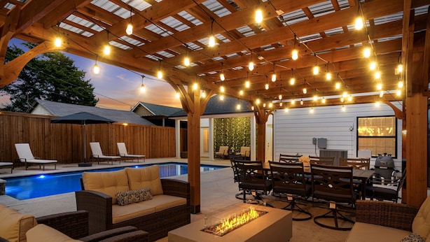 Een terras buiten met zwembad, houten pergola, lichtslingers en tuinmeubilair.