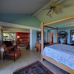 A bedroom with a four-poster bed, a ceiling fan, and a view of the living area through an open door.