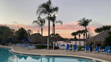 A pool area with lounge chairs, palm trees, and thatched-roof structures.
