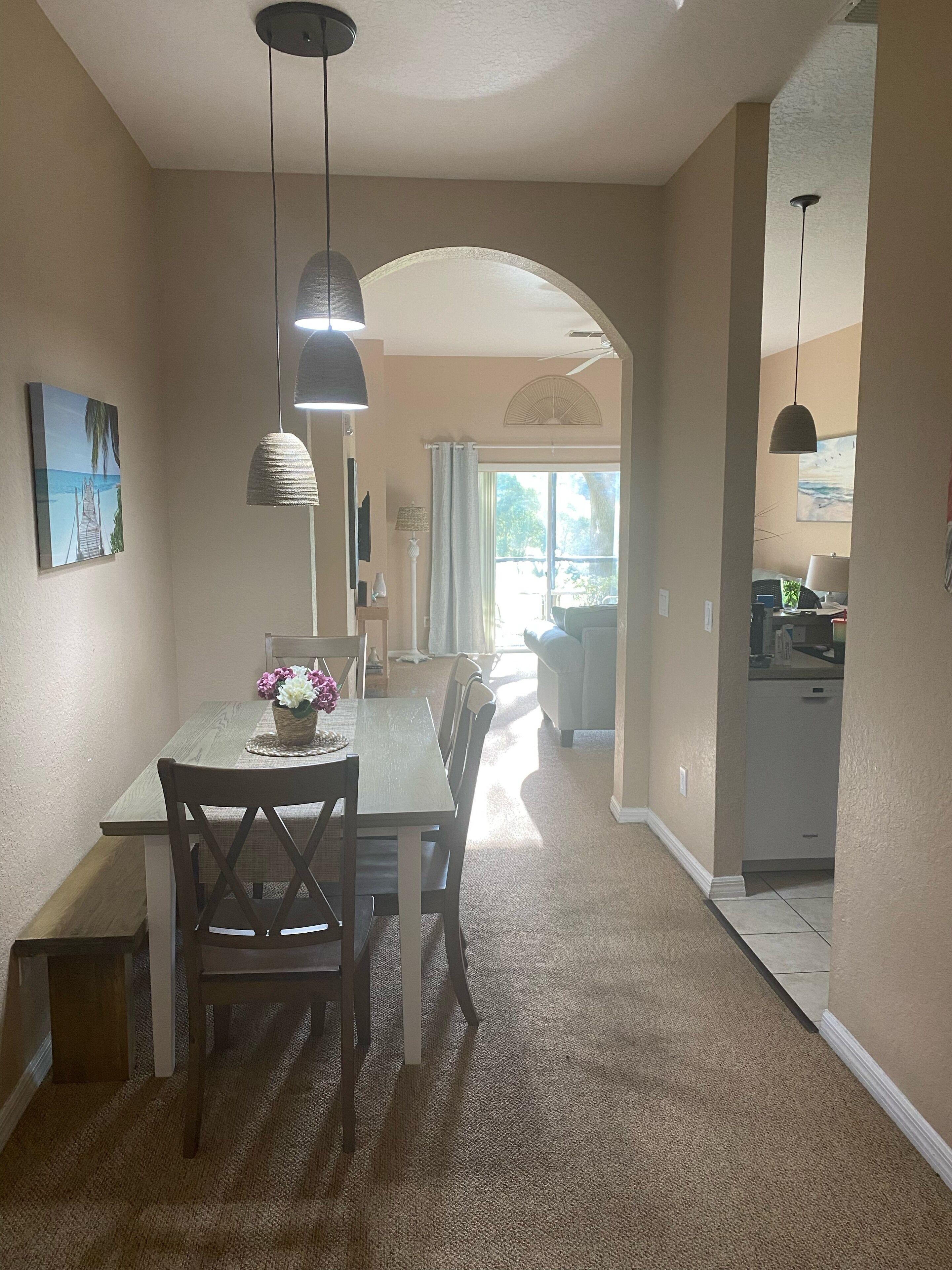 A dining area with a table and chairs, a hanging light fixture, and a view into the kitchen.