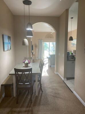 A dining area with a table and chairs, a hanging light fixture, and a view into the kitchen.