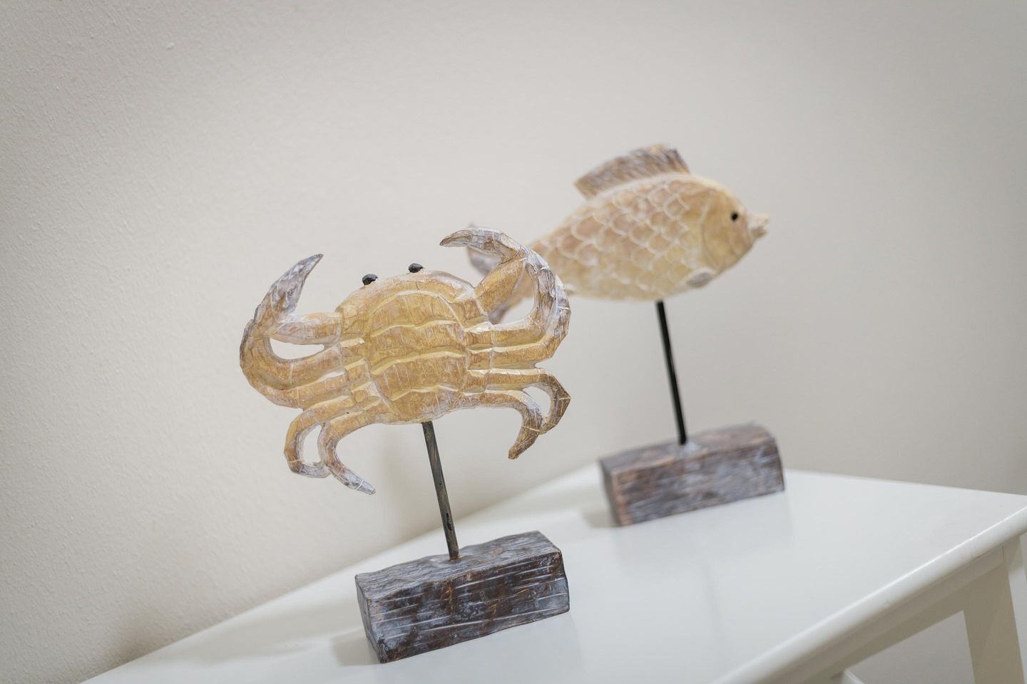 To træskulpturer, en krabbe og en fisk, står på en hvid hylde foran en ensfarvet væg.