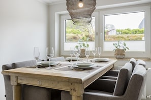 Zona pranzo con tavolo in legno apparecchiato per sei persone, lampada a sospensione in fibra naturale e due finestre con vista su piante.