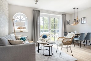 En moderne stue med sofa, rundt sofabord og stole. Der er et spiseområde med bord og stole. Store vinduer med gardiner giver naturligt lys og udsigt til det fri.