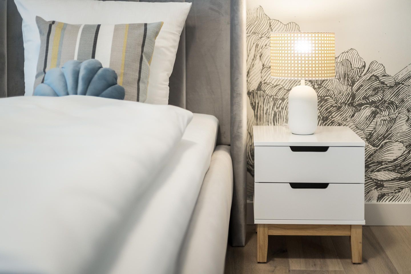En pænt redt seng med stribede puder, et natbord med en lampe, og en væg med et detaljeret mural.