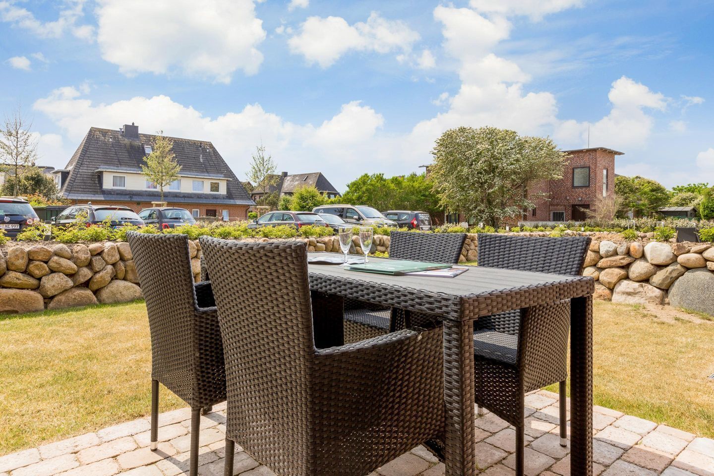 Un espace extérieur aménagé pour les repas, avec des meubles en osier, un mur en pierre et une vue sur des immeubles résidentiels et des voitures garées.