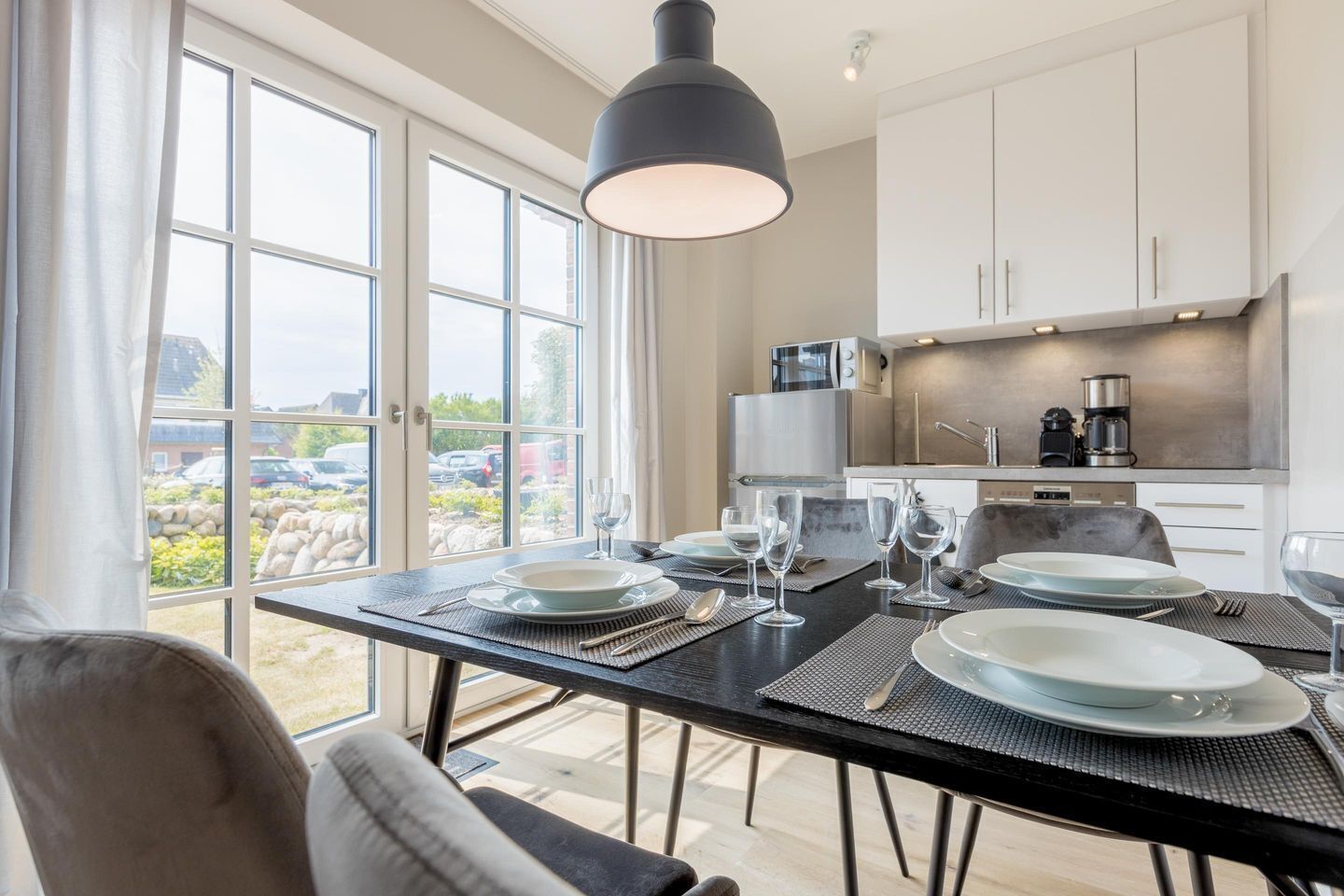 Un comedor moderno con una mesa preparada para cuatro, una cocina con electrodomésticos de acero inoxidable y vistas a un jardín a través de amplios ventanales.